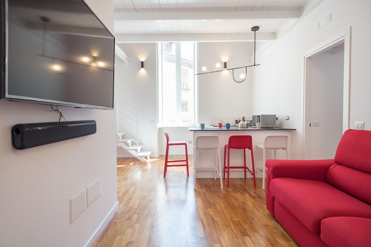 An open living area showcases a red sofa, complemented by colorful chairs around a sleek kitchen island. A wall-mounted television is positioned for easy viewing, while large windows invite natural light into the space. Bright wooden flooring adds warmth to the modern decor.