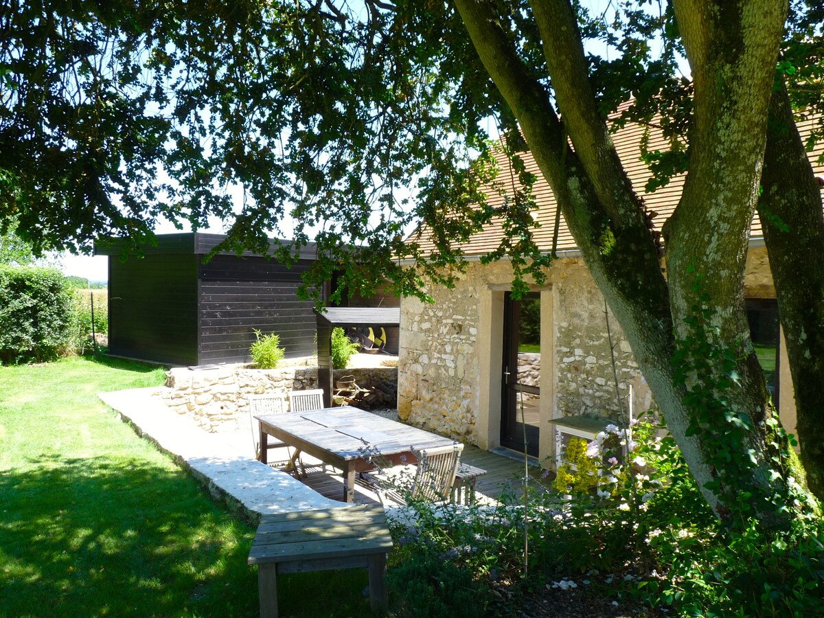 An outdoor patio area is shown, featuring a stone table surrounded by wooden seating. Lush green grass surrounds the space, with a large tree providing shade. A rustic stone and timber building is visible in the background, enhancing the serene rural setting.
