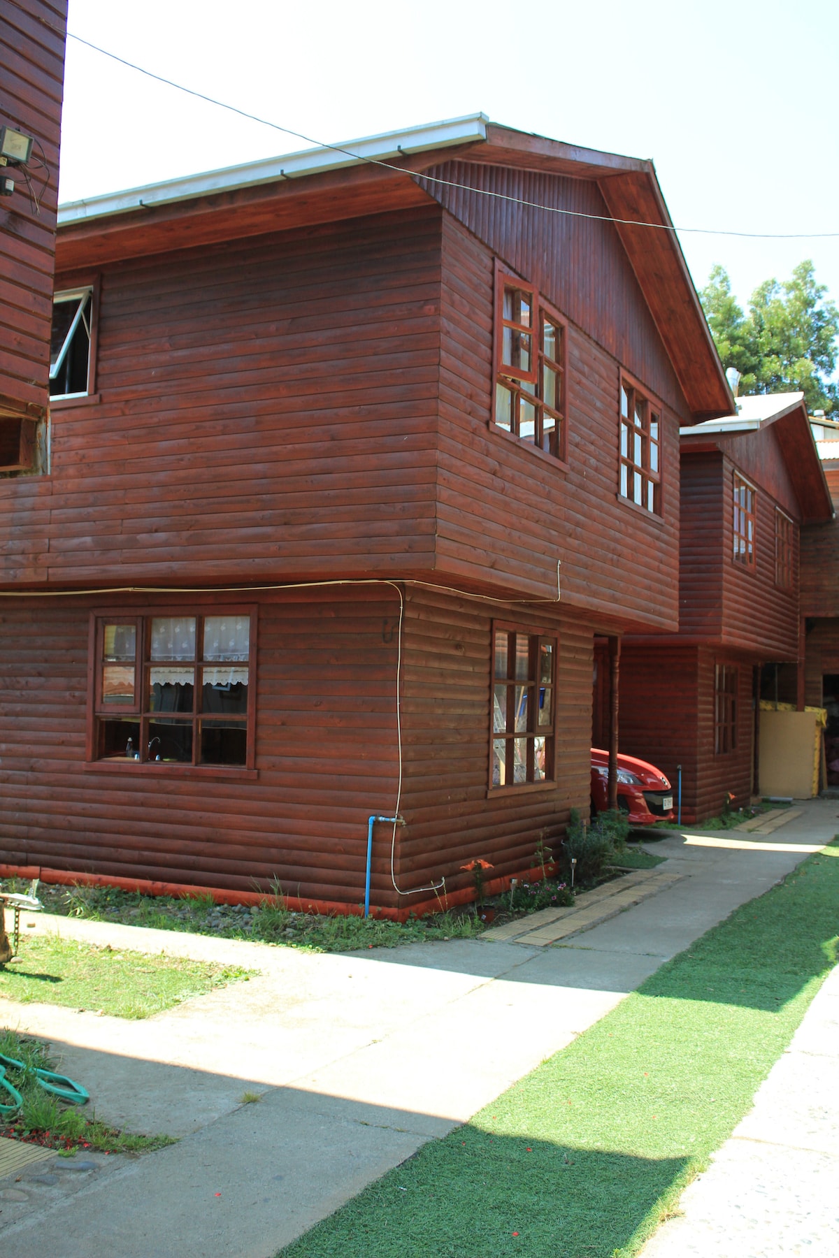 A two-story wooden cabin is depicted, featuring a rich brown exterior with multiple windows that allow natural light to enter. A well-kept pathway is visible, flanked by green grass and tidy landscaping, leading to the entrance. An adjacent vehicle is parked nearby.