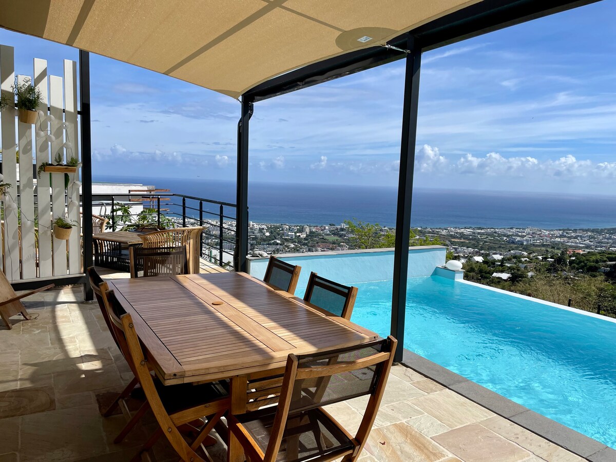 An outdoor dining area features a large wooden table surrounded by matching chairs, shaded by a canopy. The infinity pool extends towards the horizon, complemented by stunning views of the coastline and the city below.