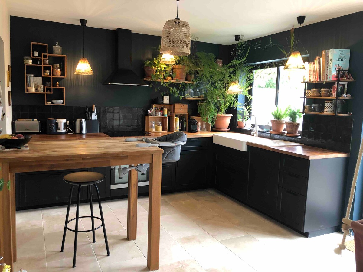 The kitchen features dark cabinetry complemented by wooden countertops. Shelves hold a variety of potted plants, enhancing the space's natural feel. Two pendant lights hang above the table, and a large window offers ample natural light, reflecting off the smooth tiled floor.