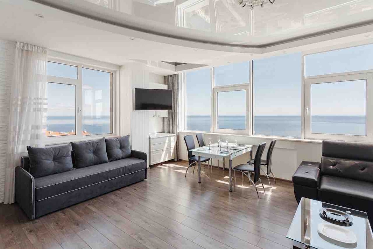 A bright living area features large windows offering an uninterrupted view of the sea. Modern furniture includes a dark sofa and a glass-top dining table surrounded by sleek chairs. A television is mounted on the wall, and soft curtains add a touch of elegance.