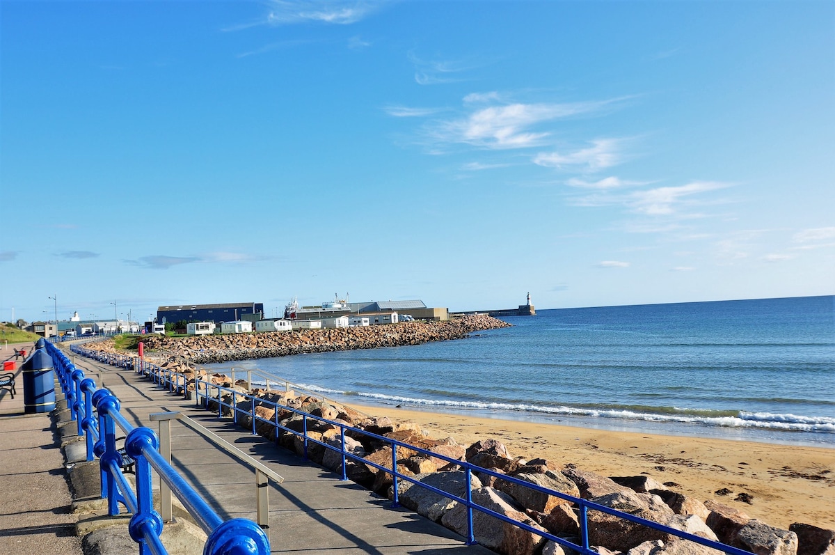 A sandy beach is bordered by a stone walkway, lined with blue railings. Gentle waves lap at the shore, and a lighthouse is visible in the distance. The sky is clear with a few clouds, creating a serene coastal atmosphere.