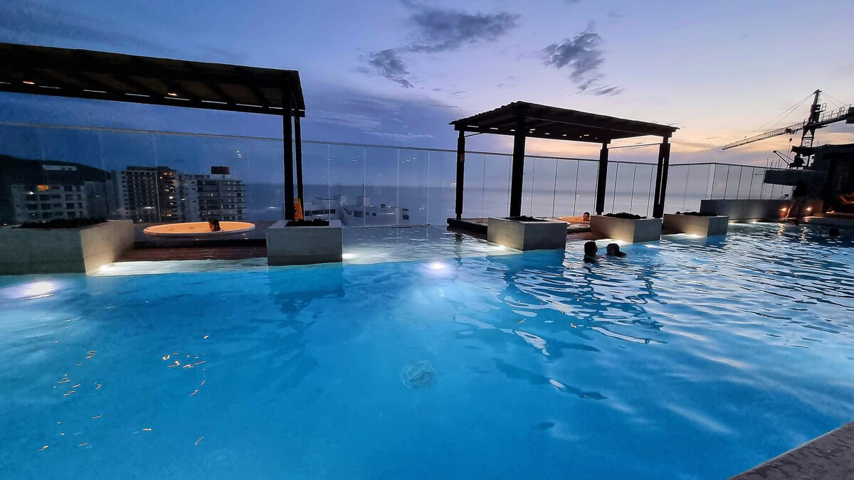 A rooftop pool shines under twilight, with gentle lighting illuminating its water. Cabana-like structures frame the pool area, providing shaded seating. The expansive horizon features a serene view of the sea, with distant city lights visible against the evening sky.