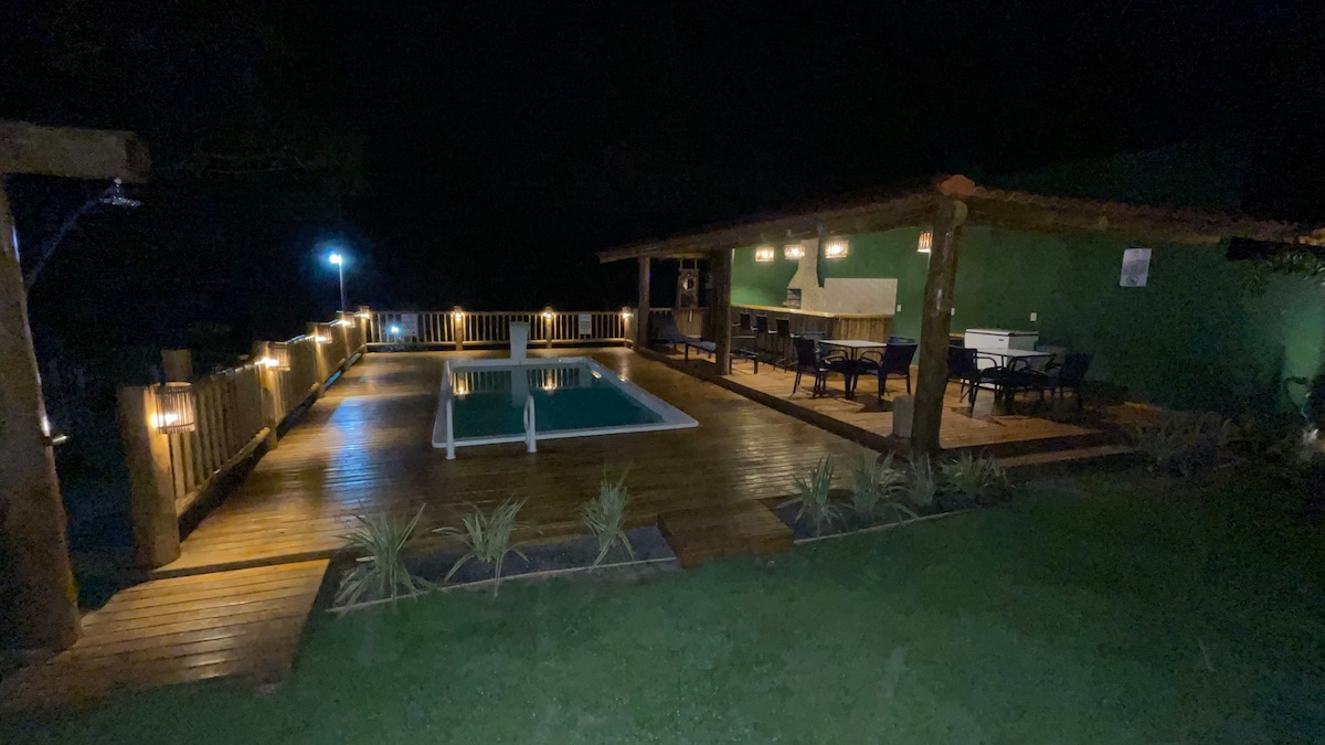 An outdoor area is revealed at night, featuring a rectangular pool surrounded by wooden decking. A covered bar area, equipped with seating and lighting fixtures, is visible. Soft lighting enhances the space, which is bordered by greenery, creating a relaxing environment.