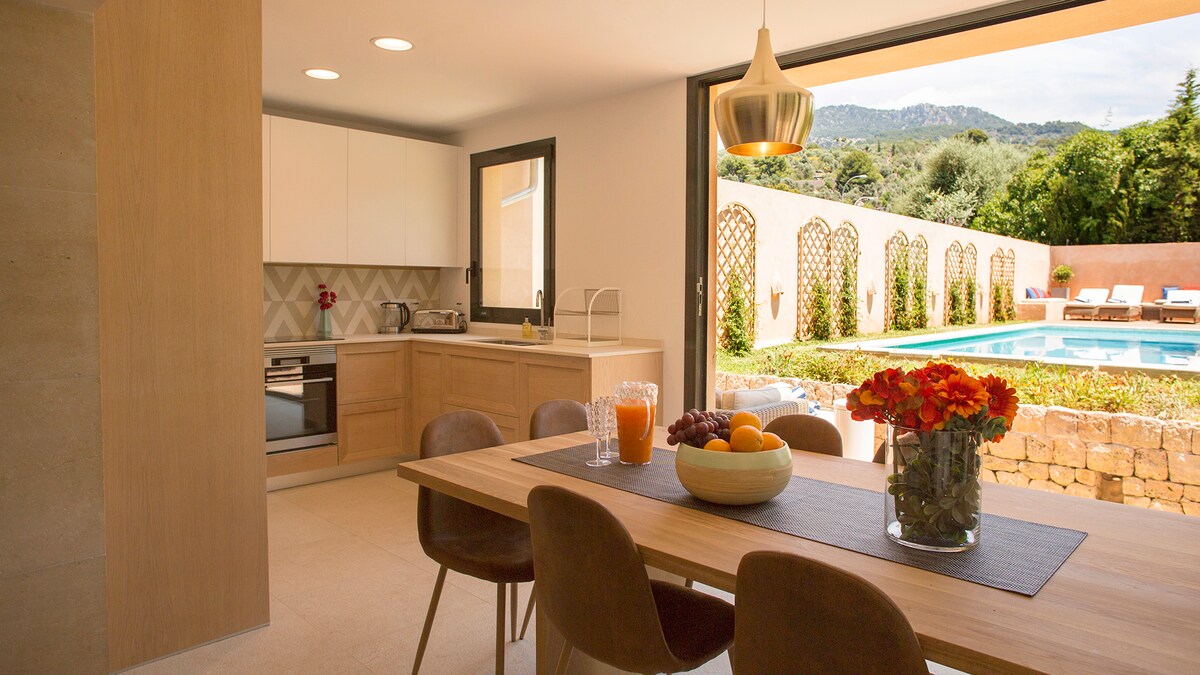 A modern kitchen area features wooden cabinetry and an open dining space with a table surrounded by four chairs. Large glass doors provide views of the pool and garden, allowing natural light to fill the room. A vase of flowers and a bowl of fruit are positioned on the table.