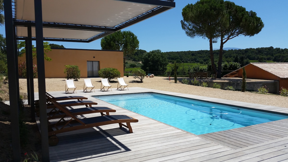 A contemporary pool area is highlighted, featuring a heated swimming pool surrounded by wooden decking. Eight sun loungers are positioned for relaxation. A covered terrace provides shade, and the expansive grounds with olive trees and distant hills are visible in the background.