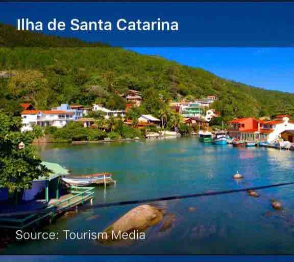 A scenic view of the coastline of Ilha de Santa Catarina, featuring colorful buildings nestled along the hills. The clear blue waters are dotted with boats, and lush green vegetation covers the surrounding landscape.