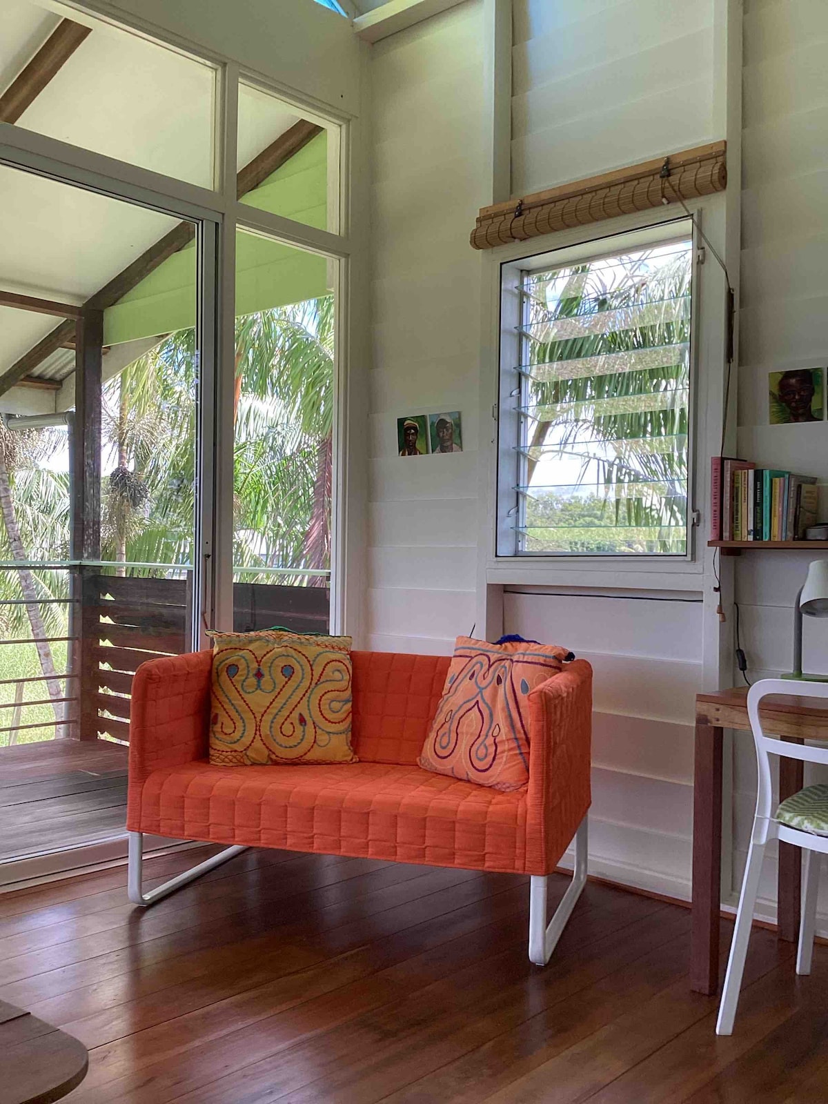 A vibrant orange couch with decorative pillows is positioned beside large glass doors, allowing ample natural light to enter the space. A wooden desk is visible nearby, along with a mirror reflecting the surroundings. Lush greenery can be seen through the open doorway.