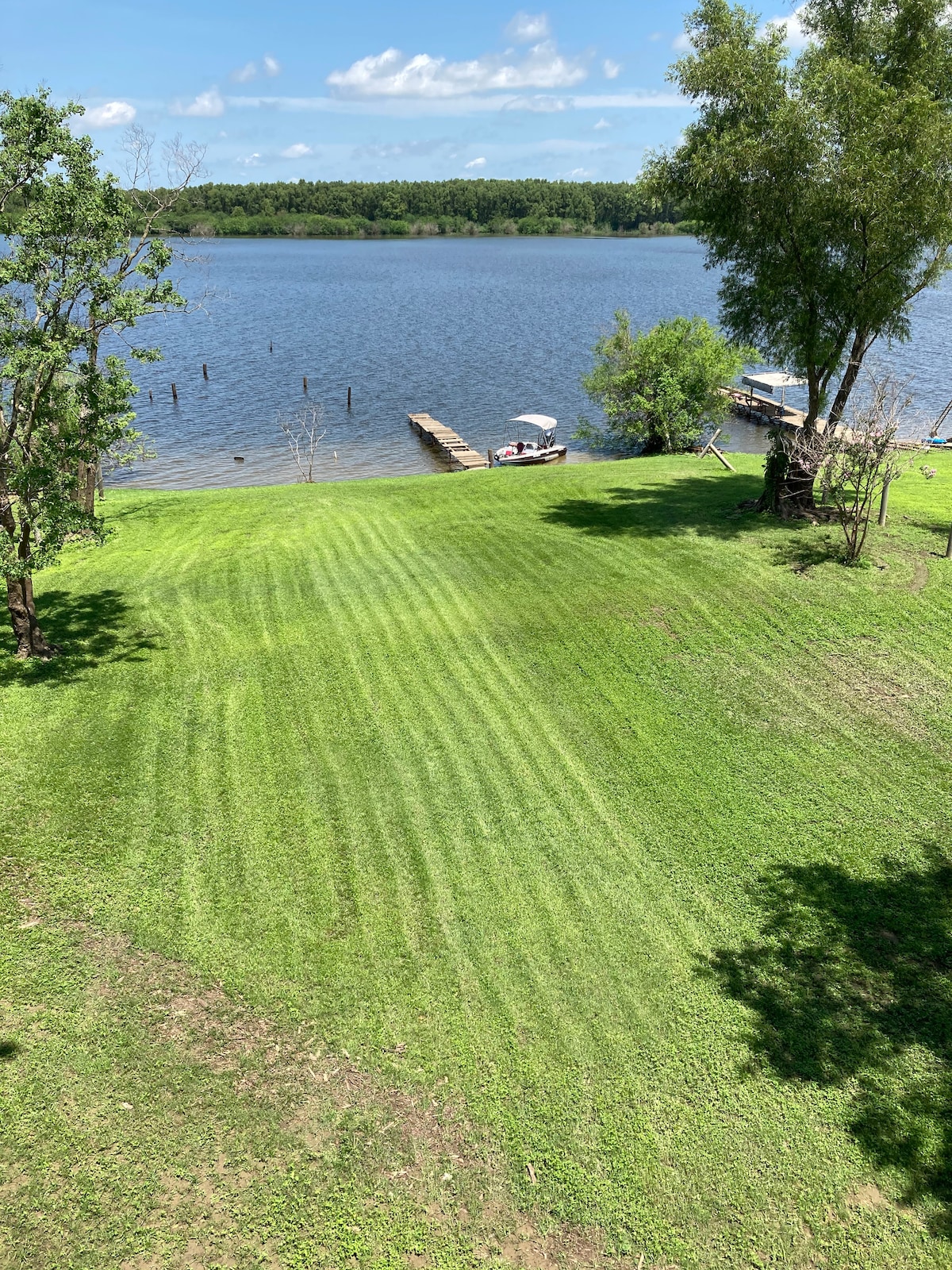 An expansive green lawn extends down to the water's edge, bordered by trees and foliage. A wooden pier is visible nearby, providing access to the lake, where a small boat can be seen anchored. The clear blue sky reflects above the tranquil water.