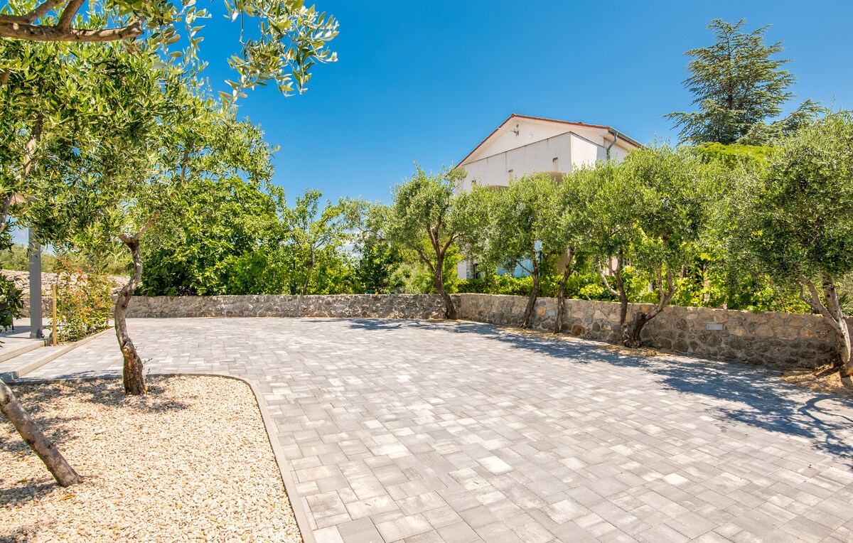 An outdoor space is visible, featuring paved stones and landscaped areas with trees, providing shaded spots. A stone wall borders the area, contributing to a serene ambiance. The clear blue sky and greenery frame the scene, enhancing the tranquil setting.