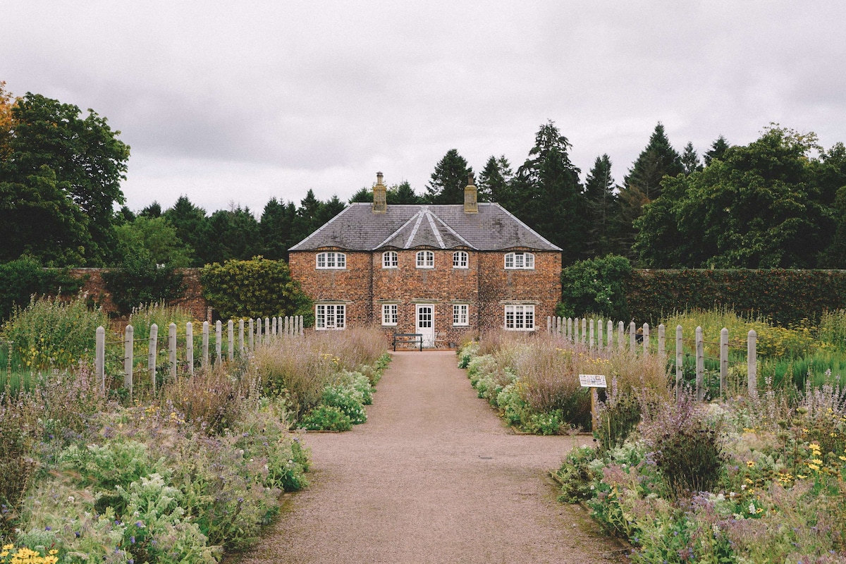 A charming 18th Century detached cottage is surrounded by lush greenery and colorful flowers. The front path leads directly to the cottage, emphasizing its inviting entrance, with a well-maintained garden framing the view. The brick façade is complemented by a traditional pitched roof and multiple windows.