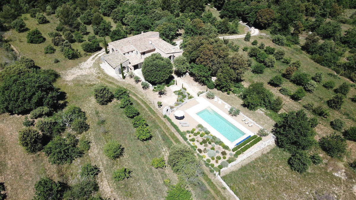 An aerial view captures the spacious property set within a lush green landscape. The vibrant garden is interspersed with trees, and the heated swimming pool is framed by comfortable loungers and sun umbrellas, emphasizing the tranquil outdoor setting.