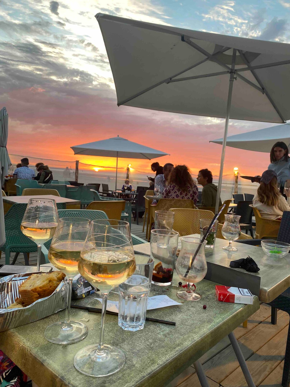 A vibrant outdoor seating area is set against a colorful sunset. Several glasses of white wine are placed on tables with a variety of snacks. Umbrellas provide shade over the diners, creating a relaxed atmosphere. The horizon showcases hues of orange and pink.