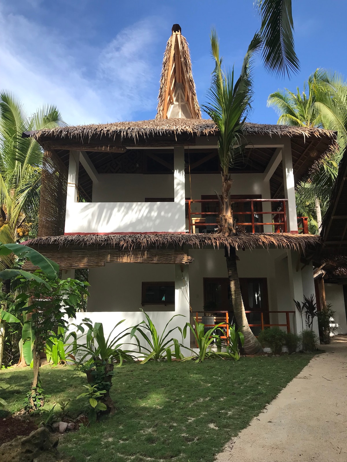 A two-story native style building showcases a thatched roof and modern architecture, surrounded by lush greenery. Palm trees and garden plants frame the property, while a pathway leads to the entrance. The structure features large openings for natural light.