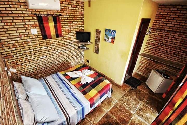 A compact room features a colorful striped bedspread covering a spacious bed, with two light blue pillows at the head. Exposed brick walls add texture, while a small television is mounted nearby. A mirror and a mini-fridge are visible, contributing to the room's functionality.