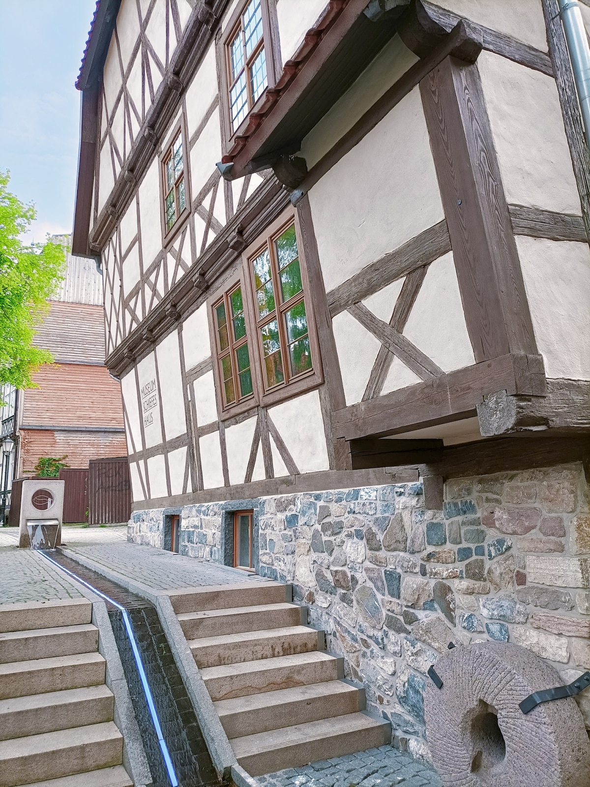 The image captures a traditional timber-framed building with a blend of white plaster and exposed wooden beams. A stone pathway leads to a staircase featuring multiple steps. Adjacent to the building, a small water feature flows, enhancing the serene environment.