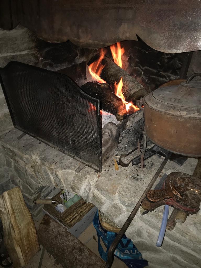 A traditional stone fireplace features a metal screen containing a vibrant fire. Logs burn brightly within, surrounded by a rustic mantel. Tools for fireplace maintenance are visible alongside neatly stacked firewood, enhancing the warm ambiance of the living space.