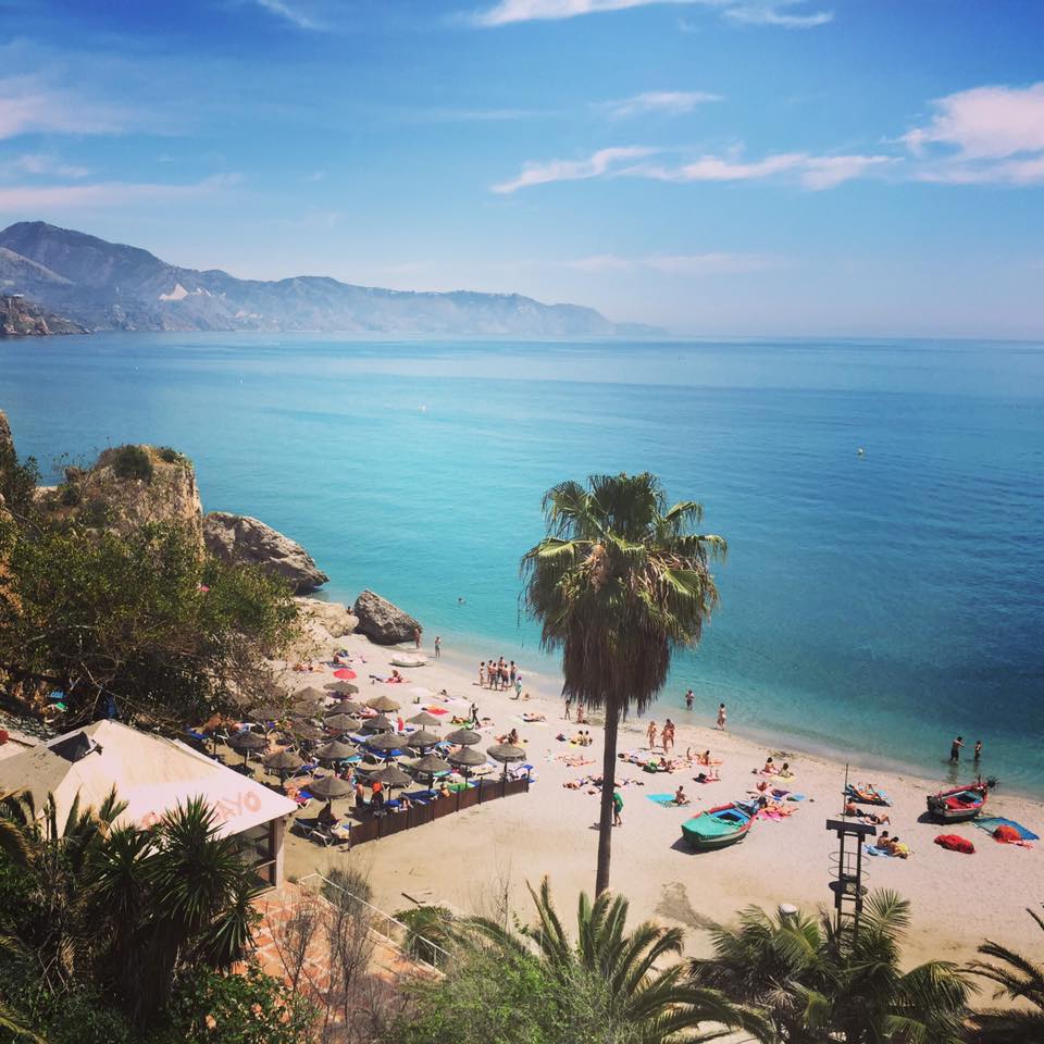 The image captures a serene beach scene with clear blue waters and gentle waves lapping at the sandy shore. Umbrellas and sunbathers populate the beach, while lush greenery and palm trees frame the foreground. In the distance, rolling hills and mountains provide a picturesque backdrop.