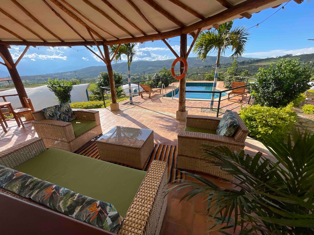 A covered outdoor seating area features comfortable wicker furniture with cushions, set against a backdrop of lush greenery and a clear view of the pool. The space is designed for relaxation, accompanied by distant mountain views and palm trees adding to the tranquil atmosphere.