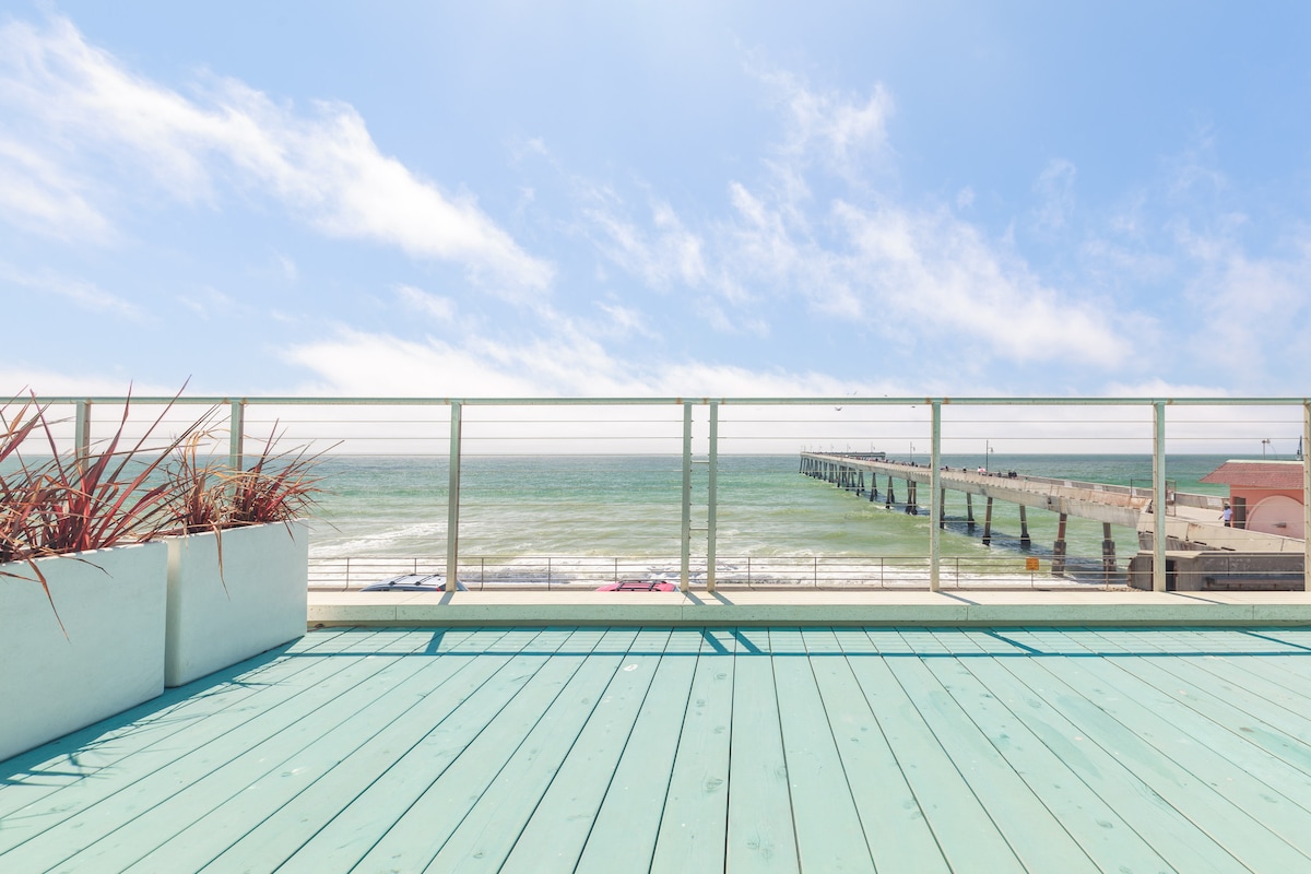 A spacious terrace features a turquoise wooden deck, overlooking the expansive Pacific Ocean and a distant pier. Decorative planters with greenery are positioned along the edge, providing a touch of natural beauty against the backdrop of the bright blue sky and shimmering water.