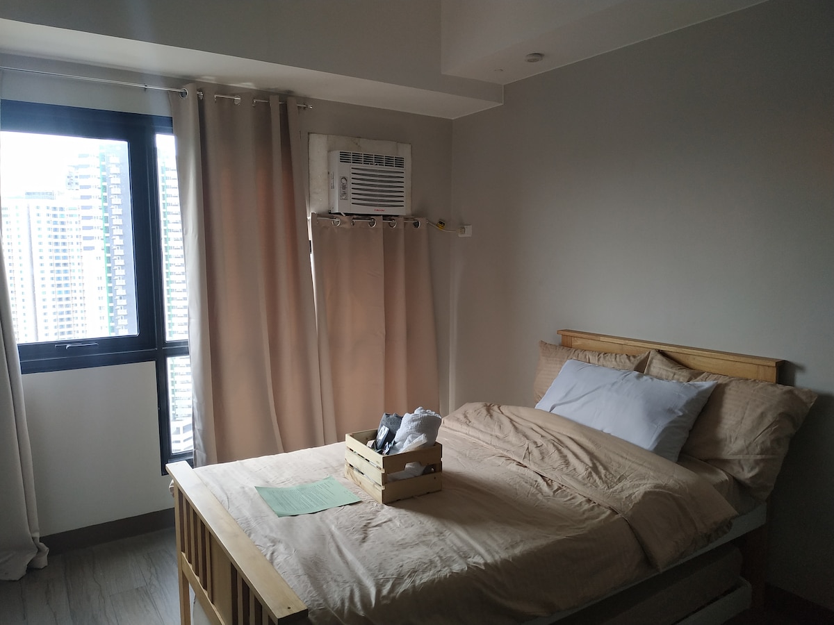A comfortable sleeping area is equipped with a spacious queen-size bed, adorned with neutral-coloured linens. Natural light filters through the large window, offering a view of the cityscape. An air-conditioning unit is mounted above the bed, enhancing the room's climate control.