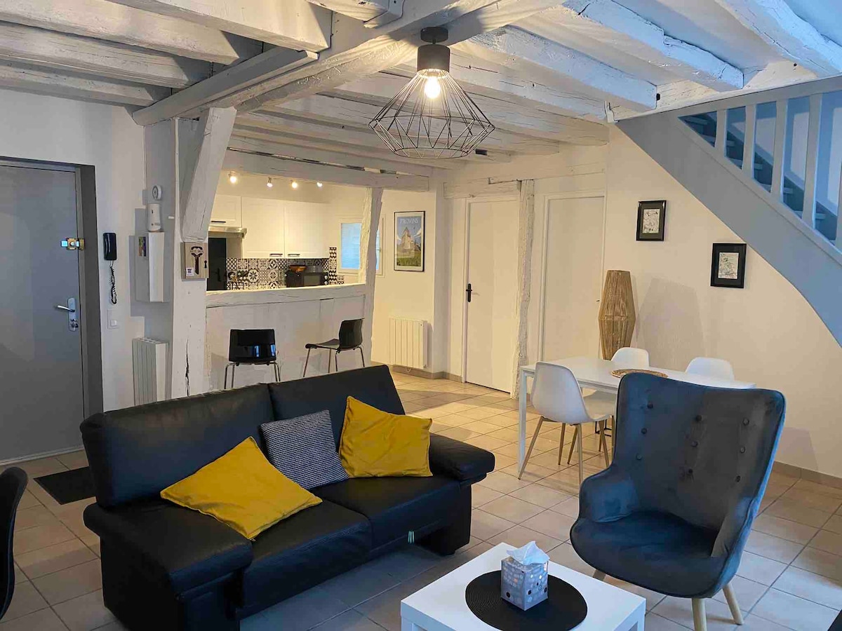 A modern living area is showcased with a dark sofa adorned with yellow cushions, complemented by a light-colored armchair. The open layout leads to a dining area featuring a white table and chairs. Exposed wooden beams enhance the space's character, illuminated by soft overhead lighting.