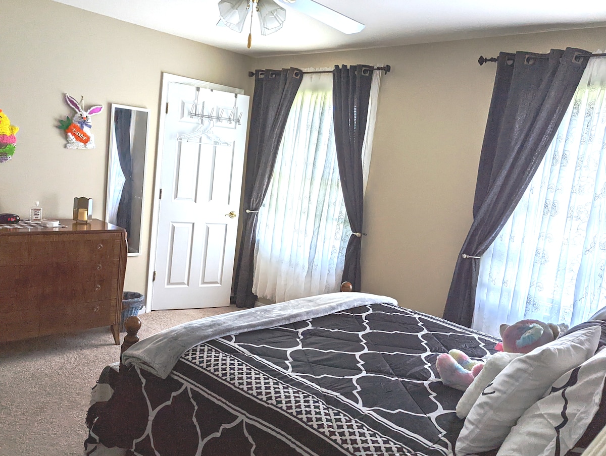 A well-lit bedroom features a spacious bed adorned with dark bedding. Sheer window treatments allow soft light to filter in. A wooden dresser stands against the wall, complemented by decorative elements, while a stuffed toy adds a touch of charm.