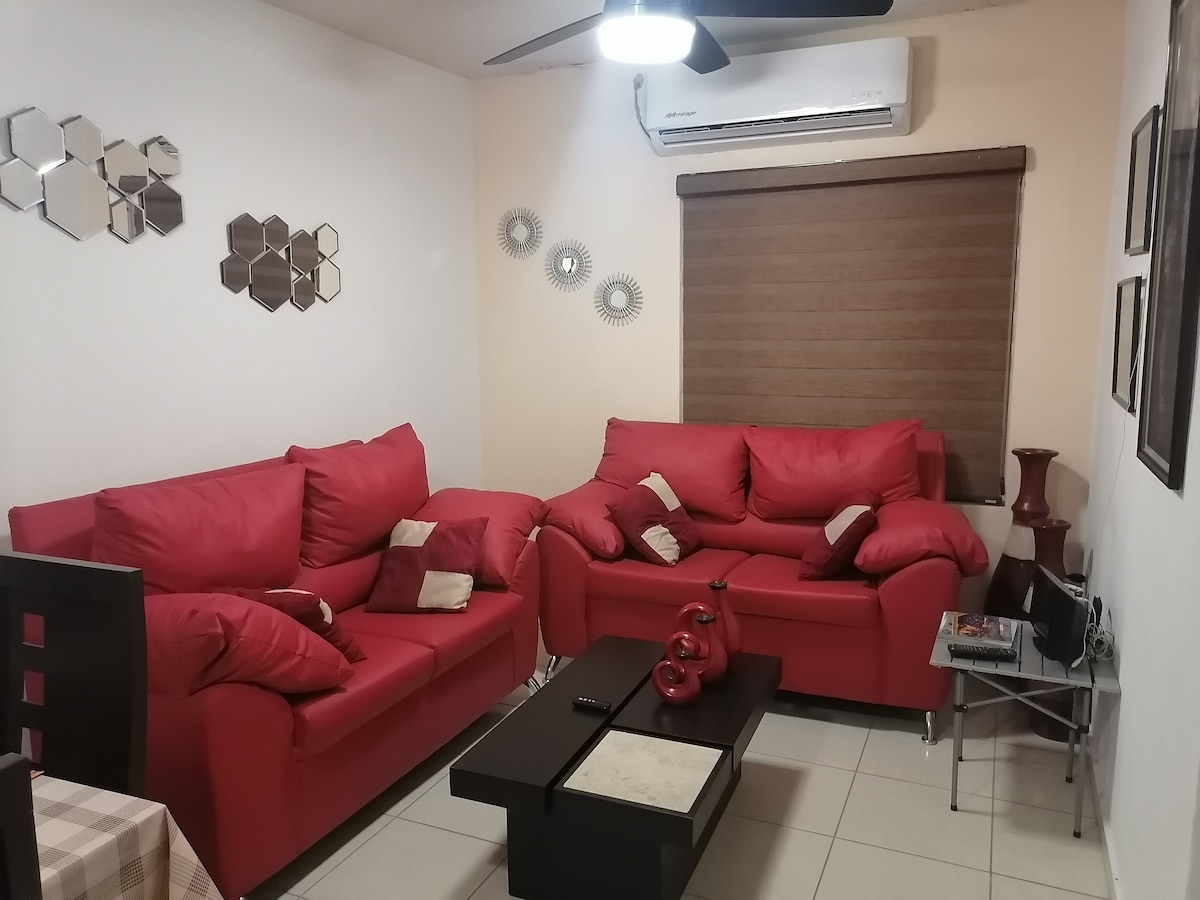 A living area features red seating arranged around a dark coffee table. Decorative mirrors are mounted on the walls, and a window is covered with a horizontal blind. An air conditioning unit is visible on the wall, contributing to the room's comfort.
