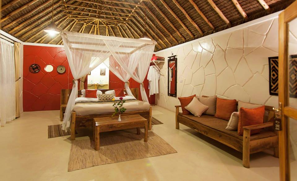 A rustic room features a thatched roof and textured walls in white and red. A spacious bed is draped with white netting, and a wooden table holds a small flower arrangement. A cozy seating area includes a wooden bench adorned with decorative cushions.