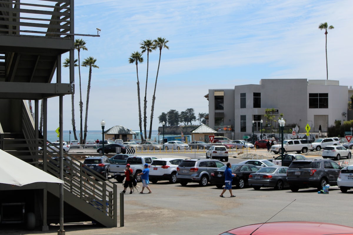 A view of a beach area showcasing a mix of palm trees and buildings. Various vehicles are parked along the waterfront, while individuals can be seen walking nearby. The ocean appears in the background, indicating proximity to the coastline.
