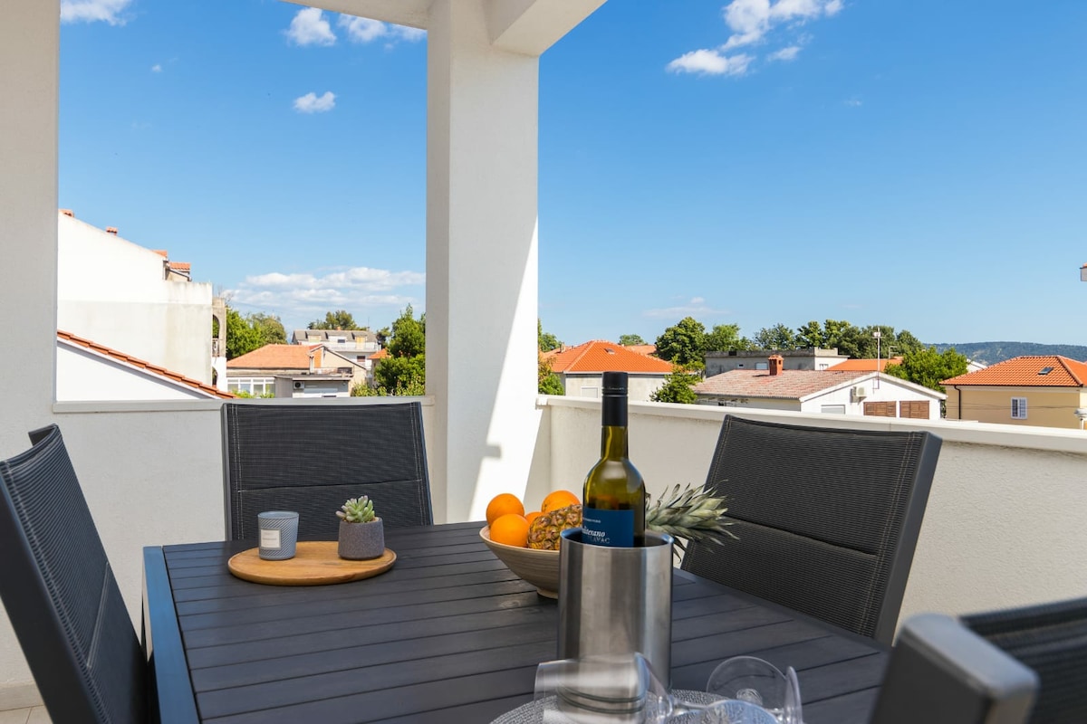 A balcony is presented with a modern dining table set for four. A bottle of wine, fruit bowl, and decorative items adorn the table. Clear blue skies and rooftops of nearby buildings are visible in the background, contributing to a tranquil outdoor space.