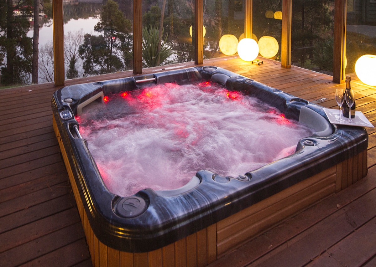 A spacious jet spa is positioned on a wooden deck, surrounded by large windows offering views of the lake. Soft red lights illuminate the water, creating a calming ambiance. A bottle of sparkling wine rests nearby, inviting guests to relax and unwind.