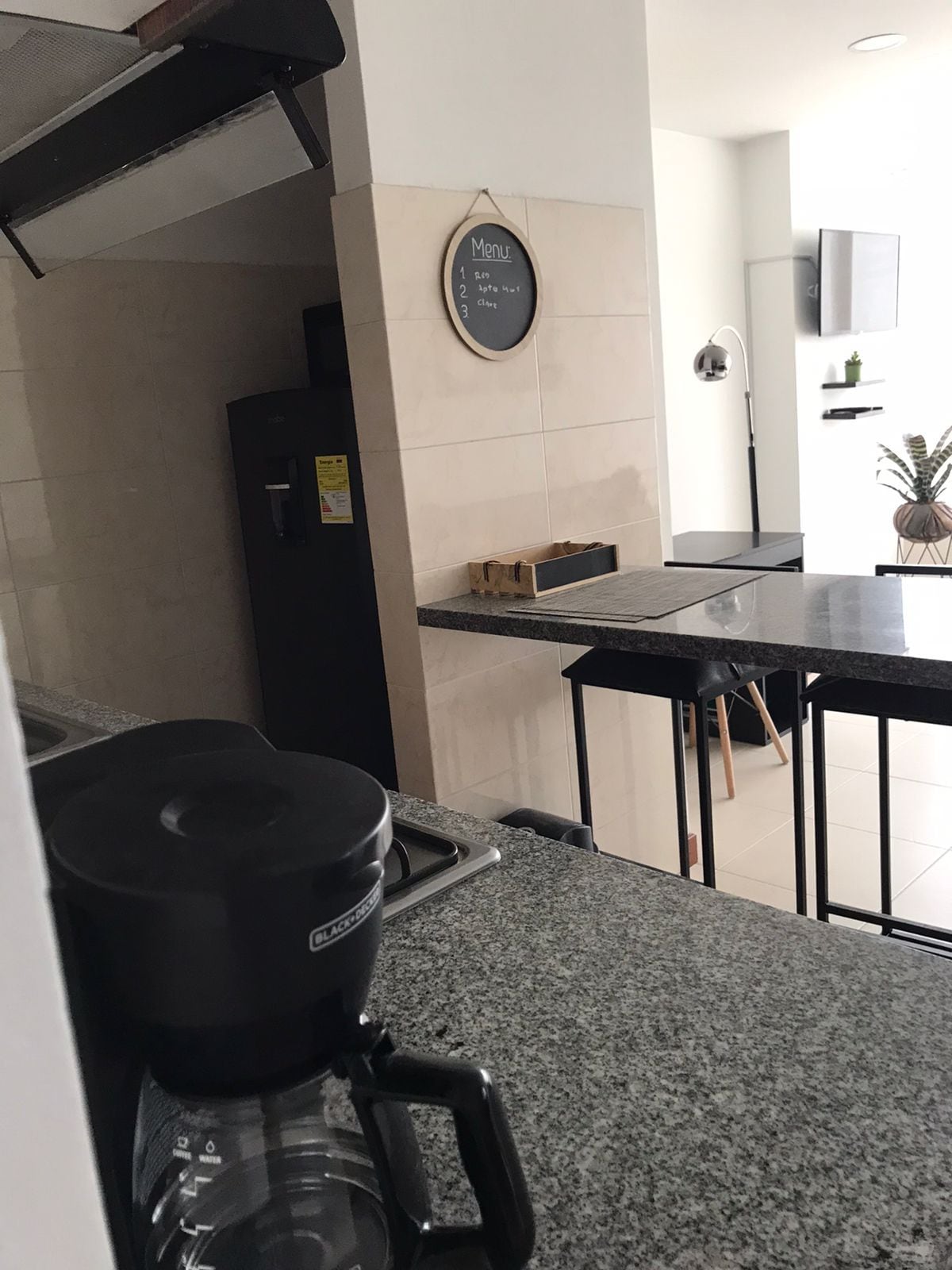 The kitchen showcases a sleek granite countertop with a coffee maker prominently displayed. A dark refrigerator is positioned nearby, and a chalkboard adds a personal touch to the wall. A dining area is visible in the background, featuring a modern table and chair setup.