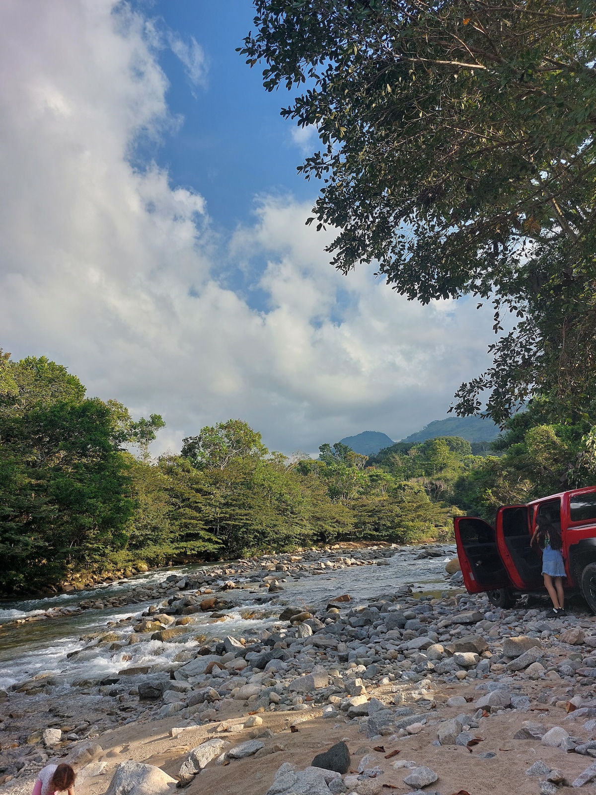 A serene riverbed is surrounded by lush greenery and distant mountains. Large rocks and pebbles are visible along the water's edge, while a red vehicle is parked nearby. The sky displays a mix of clouds and blue, suggesting a pleasant day in nature.
