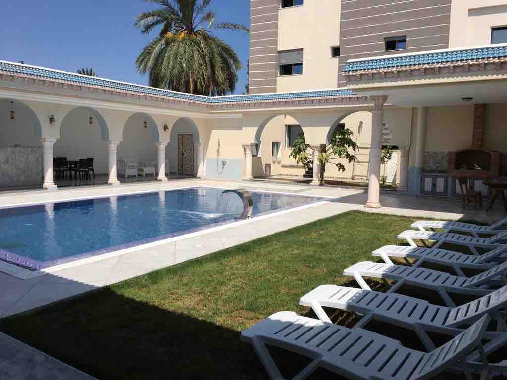 A private swimming pool is surrounded by manicured grass and lounge chairs. Archways lead to a spacious area featuring palm trees and a light-colored building in the background. The bright blue sky enhances the inviting atmosphere, creating a serene setting for relaxation.