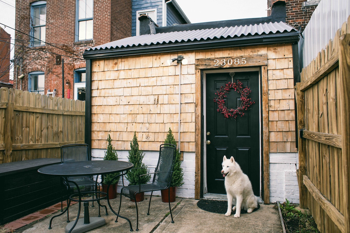 The exterior of the cottage is showcased with wooden shingles and a festive wreath on the door. A white dog sits near the entrance, while a small patio area features a table and chairs surrounded by potted evergreen plants and a fenced yard.