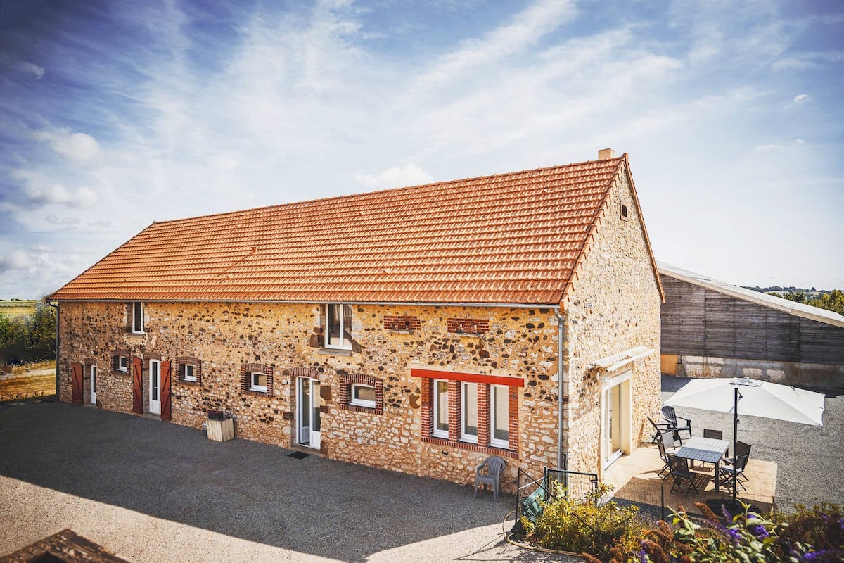 A rustic stone building with a gabled red-tiled roof is shown, surrounded by a gravel area. Numerous windows and doors add character to the facade, and outdoor seating is visible, providing space for relaxation. The backdrop consists of a clear blue sky with scattered clouds.