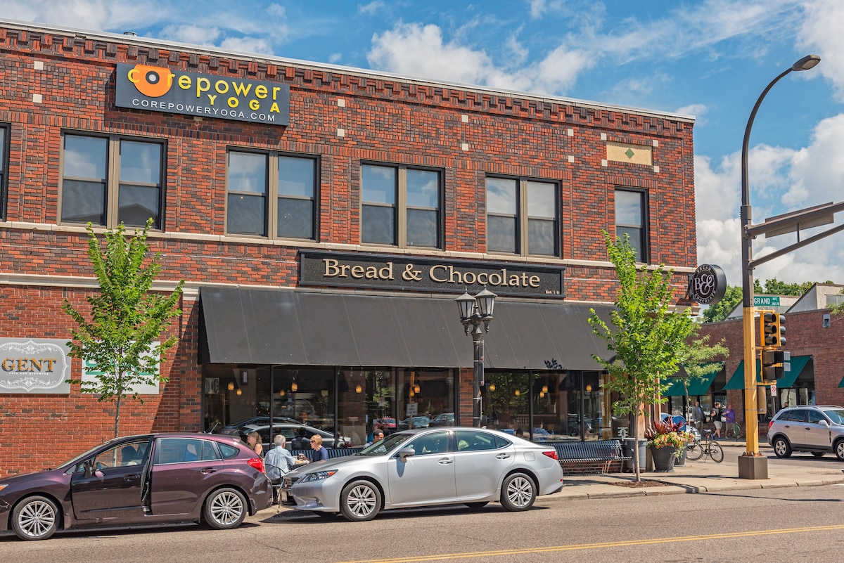 A brick building houses a café named 'Bread & Chocolate' visible under a wide awning. Outdoor seating is present along the sidewalk, with patrons engaging at tables. Green trees line the street, enhancing the lively urban scene.
