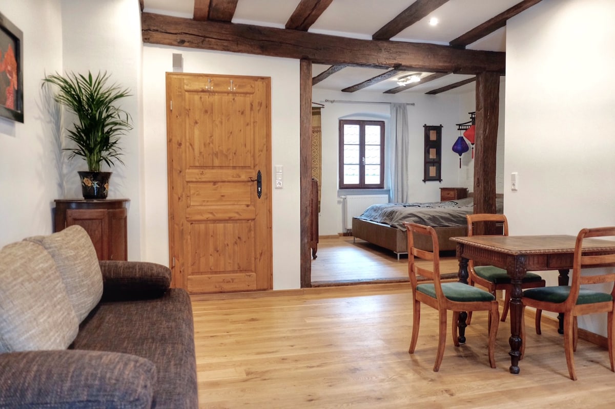 A cozy living area features a brown sofa and a wooden dining table with four chairs. Exposed beams add character to the room. A separate sleeping area is visible through an archway, showcasing a spacious bed and natural light streaming from a nearby window.