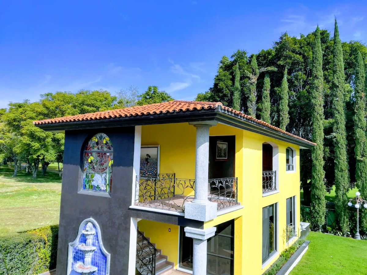A vibrant yellow house features a red tile roof and decorative details. Balconies provide views of the surrounding greenery and golf course. Towering trees frame the property, enhancing the peaceful outdoor setting.