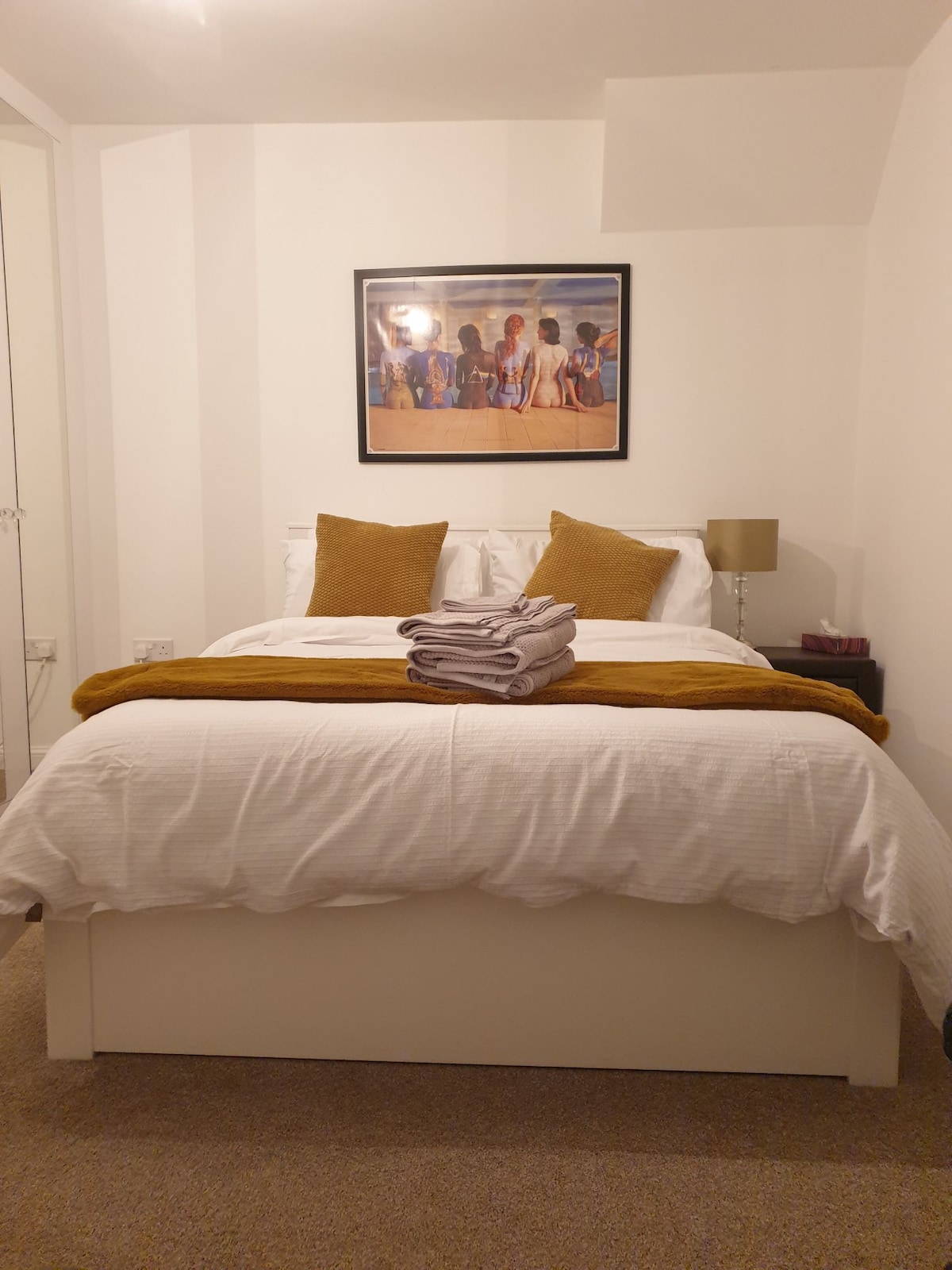 A well-appointed sleeping area is present, featuring a spacious bed dressed in white linens, complemented by mustard-colored cushions. A folded towel is placed at the foot of the bed. A framed artwork hangs above the headboard, with a bedside lamp providing soft illumination.