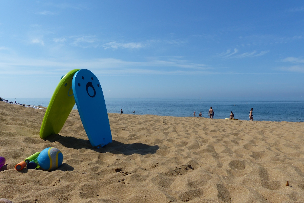 A sandy beach is highlighted, with gentle waves lapping at the shore. A blue and yellow beach chair stands in the foreground, while beachgoers can be seen in the distance. Bright sunlight fills the scene, enhancing the coastal atmosphere.