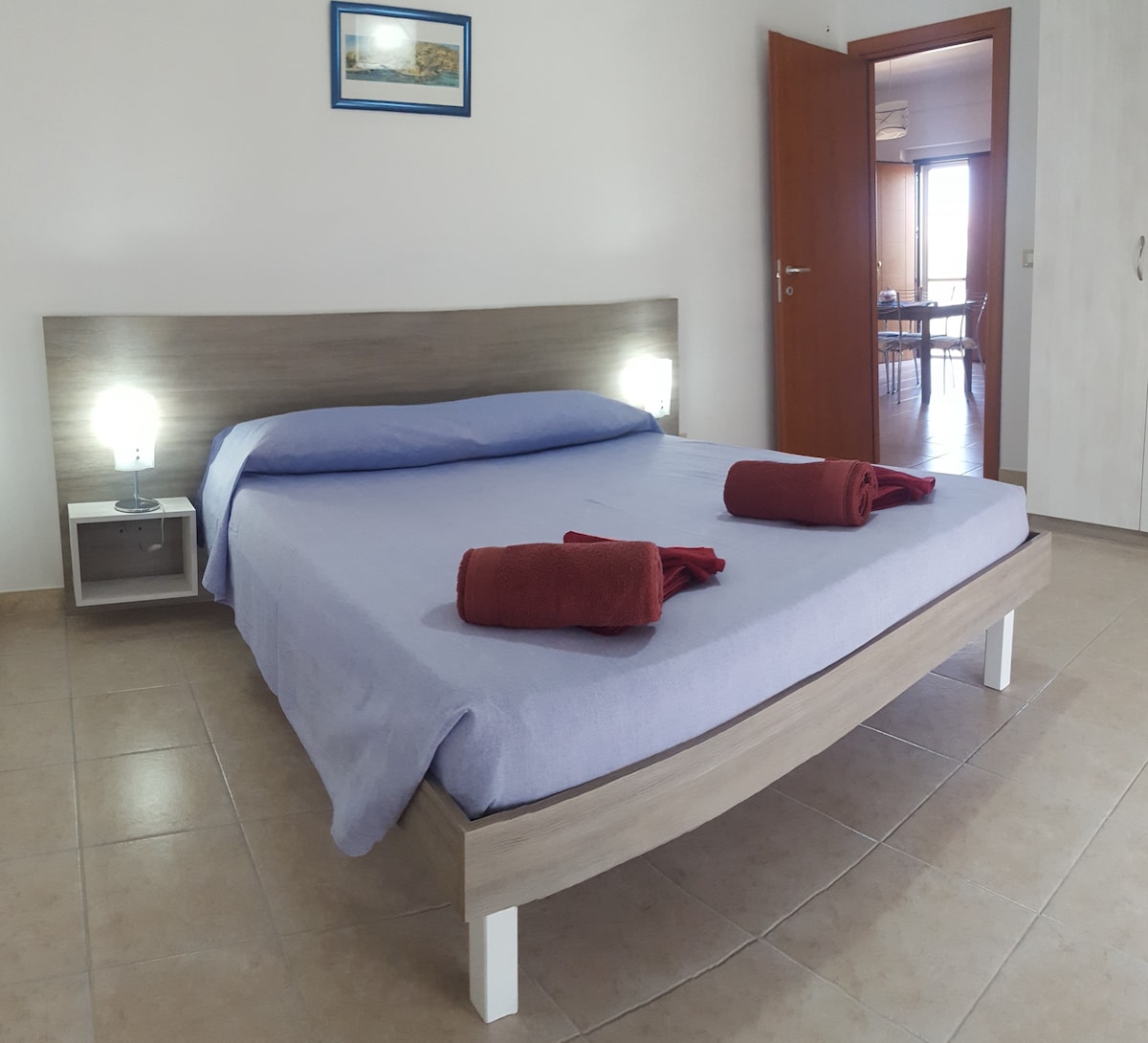 A spacious bedroom is presented with a large bed dressed in soft blue linens. Two bedside lamps provide gentle lighting while rolled towels are neatly placed at the foot of the bed. A wooden headboard complements the room's neutral tones, and a framed artwork is mounted above.