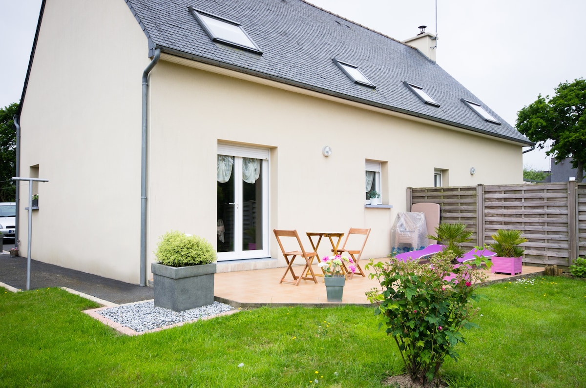 The exterior of the house features a spacious terrace, furnished with two wooden chairs and a small table, complemented by potted plants. A grassy area surrounds the terrace, providing a serene outdoor space. Large windows allow natural light to flood the interior.