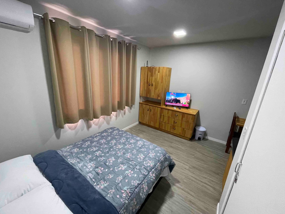 A private bedroom is presented with soft gray walls and natural light filtering through beige curtains. A spacious bed is covered with a floral quilt. A wooden cabinet and a small desk are visible, alongside a television mounted on the wall.