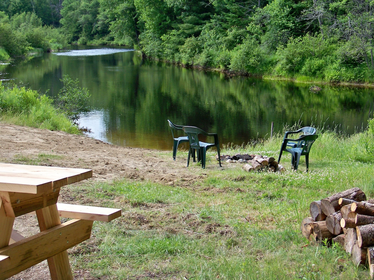 A serene riverside setting is depicted, featuring two green lawn chairs positioned near the water. A sandy area leads to the river, while a rustic picnic table is visible in the foreground. Stacked firewood sits nearby, surrounded by lush greenery.