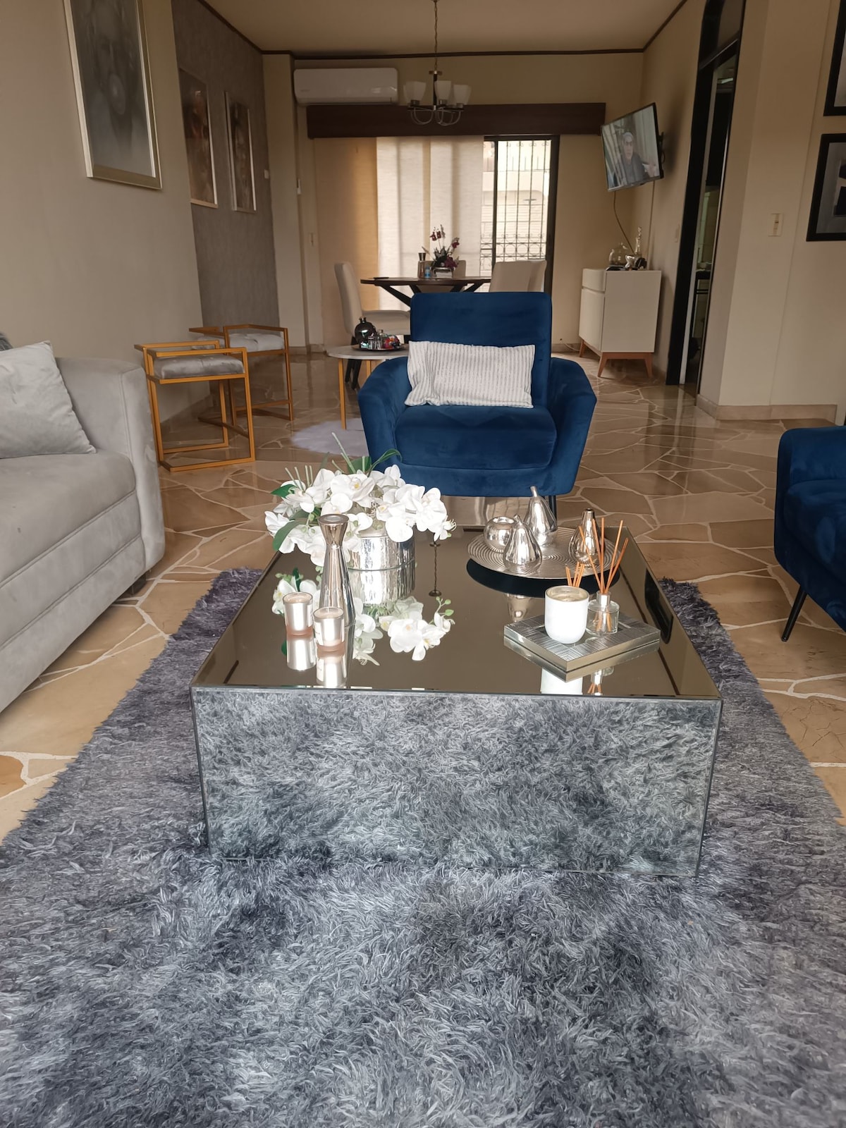 A modern living area features stylish blue chairs and a light gray sofa. A mirrored coffee table rests on a soft, textured rug adorned with decorative items including flowers and candles. Natural light enters through large windows dressed with simple shades.