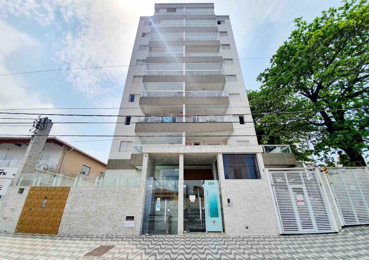 A modern multi-story building is presented, featuring a contemporary façade with large glass balconies. The entrance is framed by a sleek stone structure and an automatic gate, surrounded by greenery. Paved pathways lead up to the entrance, which is well-lit and open.