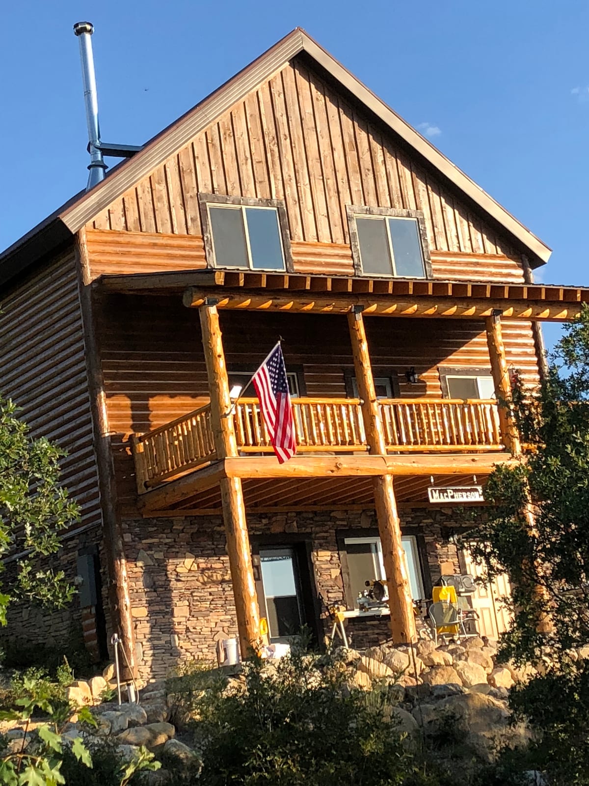 The three-story cabin features a rustic log exterior with stone accents. A wooden balcony extends from the second floor, adorned with an American flag. Large windows provide natural light, and the surrounding landscape includes greenery and a stone pathway leading to the entrance.